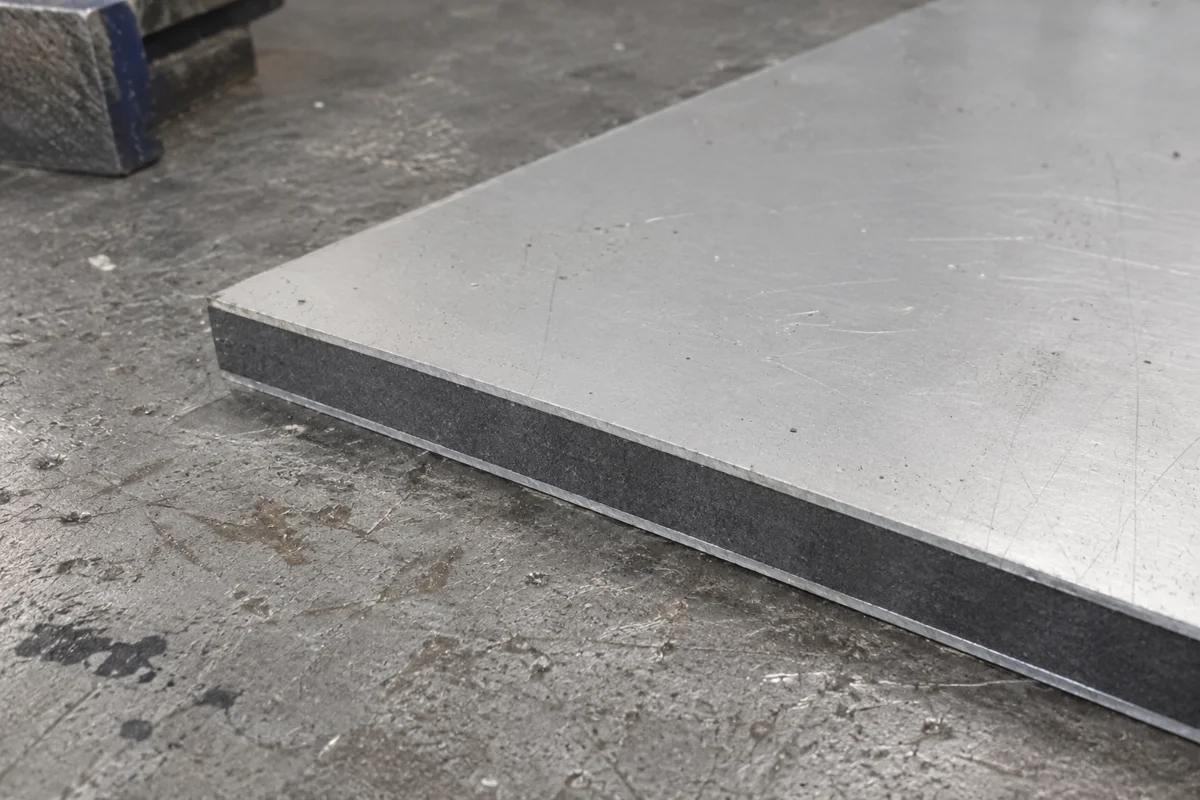 Canto de un panel con dos caras metálicas delgadas y un núcleo gris sobre una mesa de taller